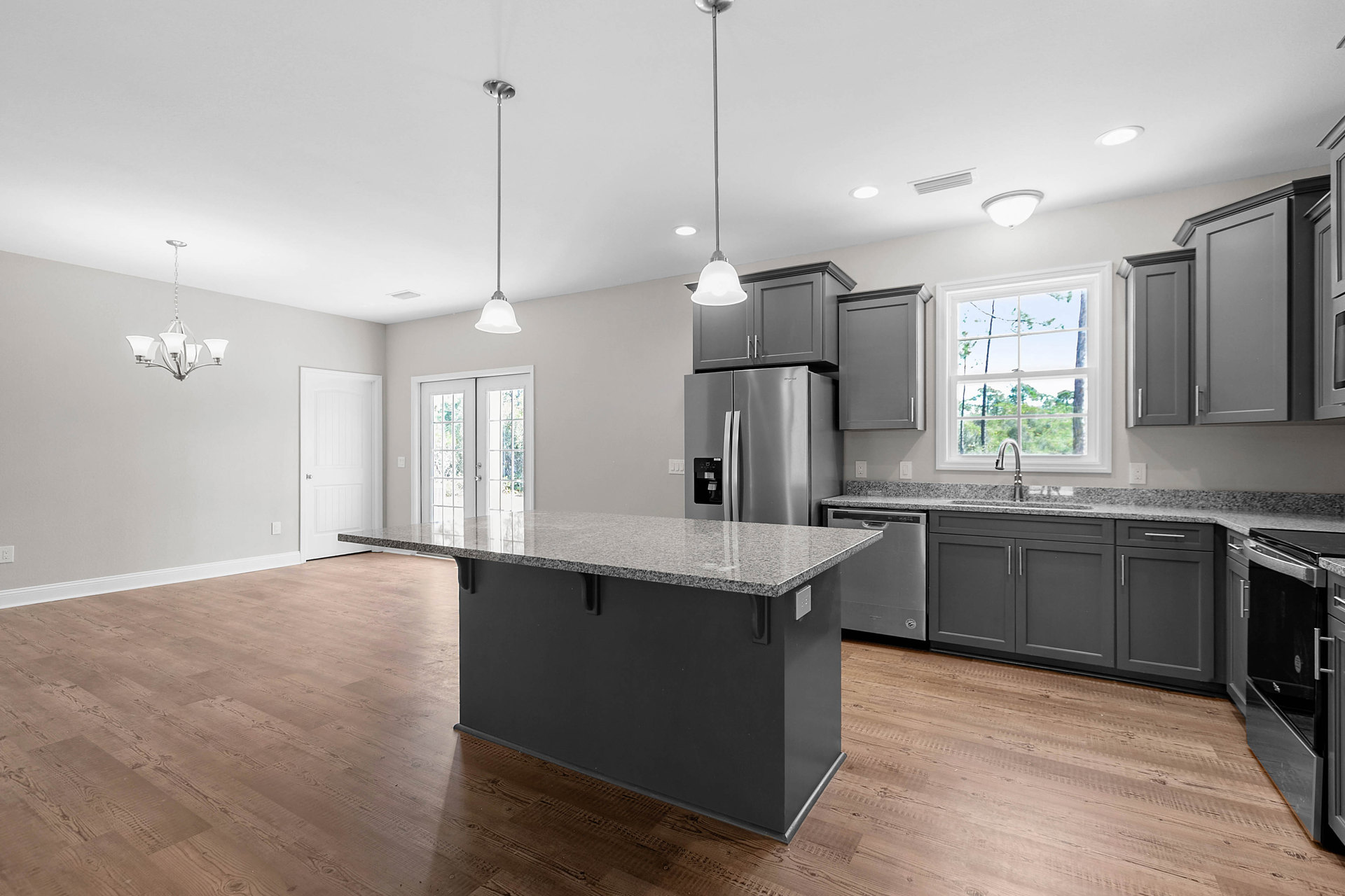 Spacious kitchen featuring a large central island with black countertop, white cabinetry, stainless steel refrigerator, window above sink overlooking trees, and white door with