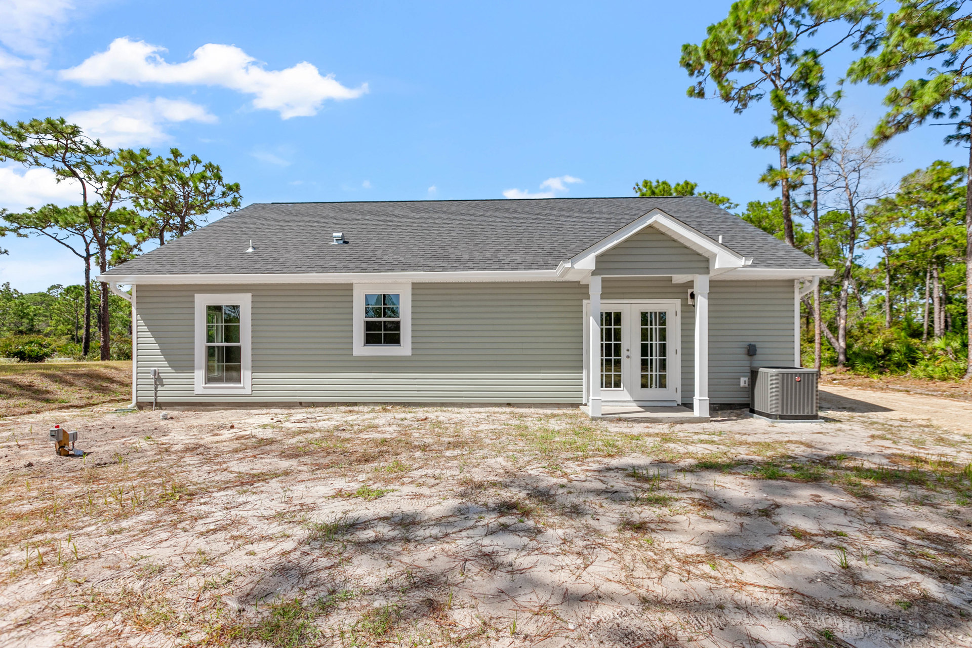 Modern cottage-style home with white siding, large window with white frame, double glass doors, dirt yard, scattered grass, mature trees, and visible outdoor air conditioner unit