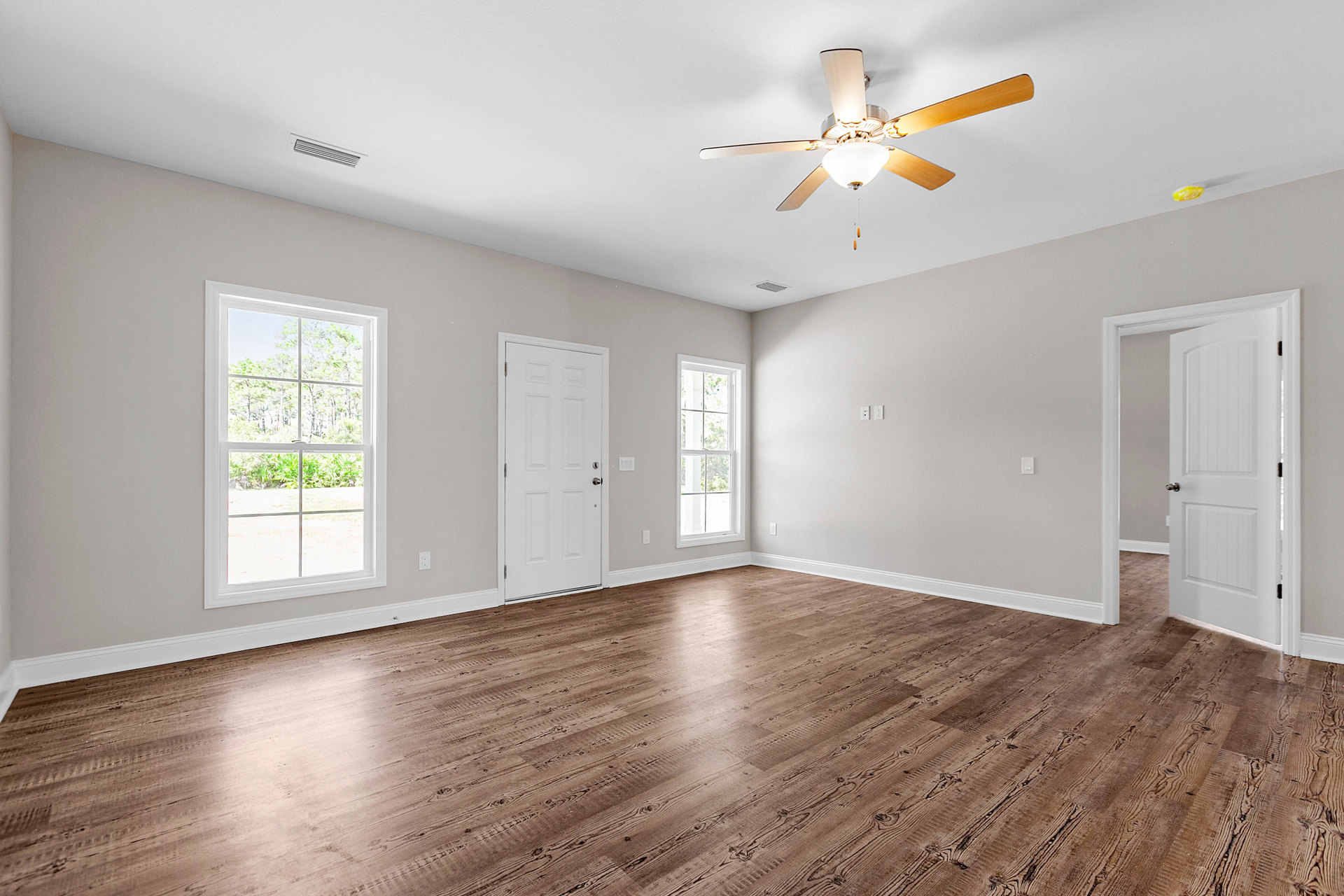 Ceiling fan with light fixture, wood flooring, white walls, window with white frame overlooking trees, white door with silver handle