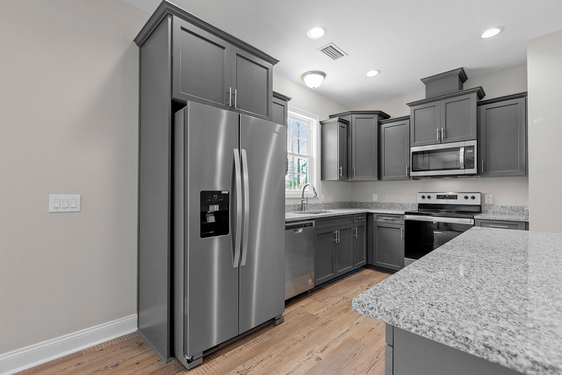 Grey shaker cabinets, stainless steel refrigerator with open door, built-in microwave, white quartz countertop, undermount sink, light switch on wall, wood flooring