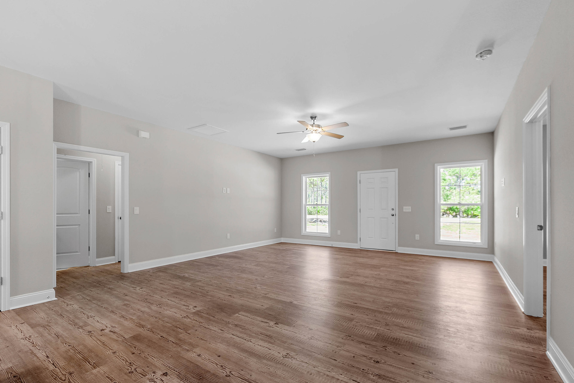 Spacious room with wood flooring, white walls, ceiling fan with light fixture, multiple windows showing trees outside, and a white door with silver handle