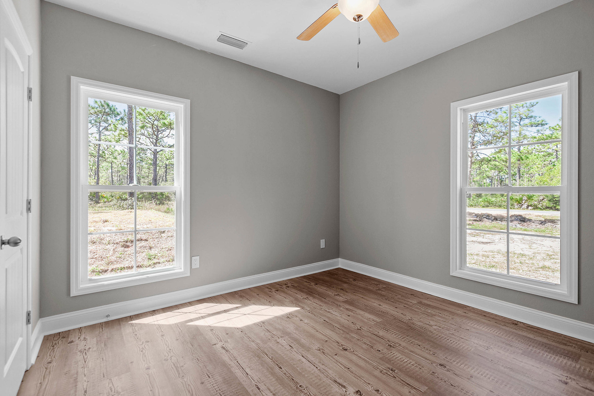 Bright room with wood flooring, white plaster ceiling, ceiling fan, large windows showing trees outside, and a white door