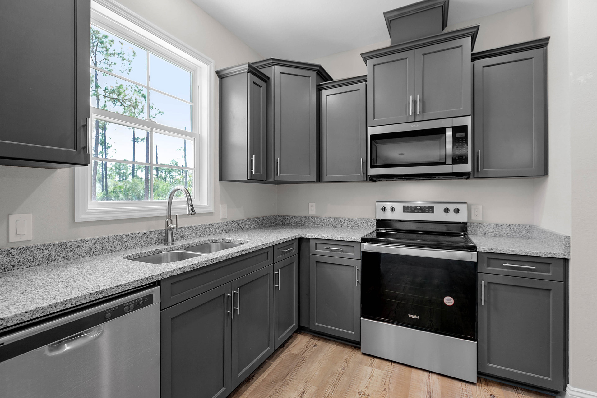 Grey shaker cabinets, black and silver stove, open microwave door, white countertop, window with trees outside, close-up of cabinet and white door