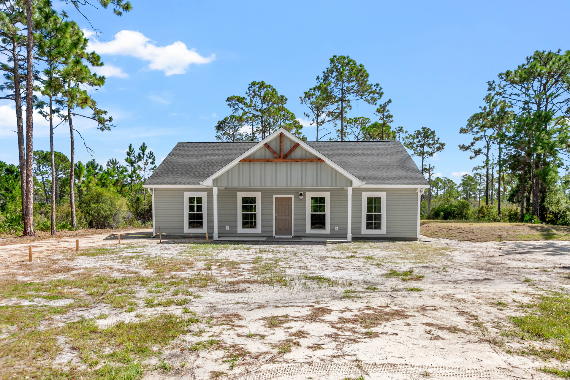 Two-story house with brown door and white trim, white-framed windows, surrounded by dirt patches and mature trees; small white cottage visible in the background.