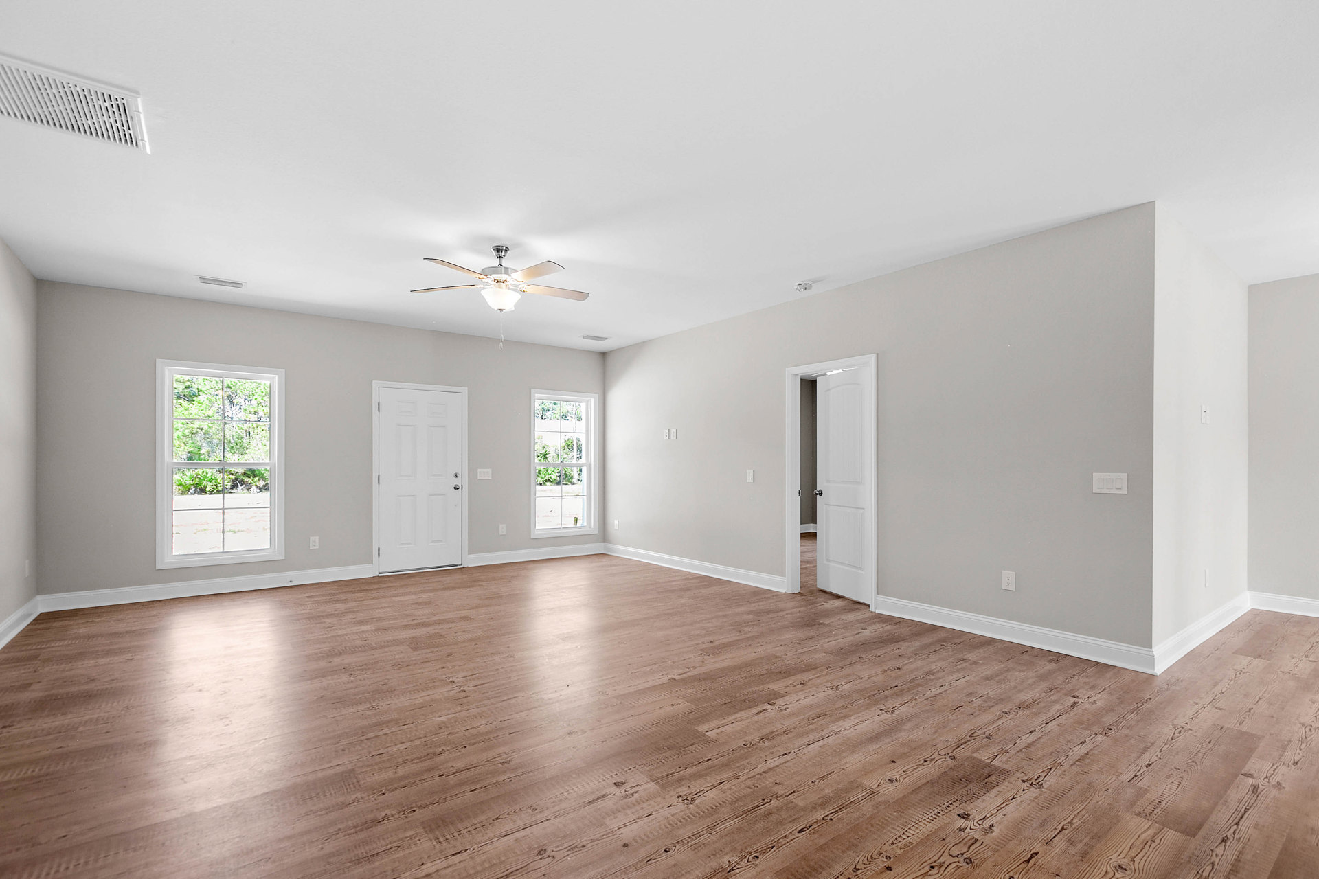 White-walled room with wood flooring, ceiling fan with light, white door with silver handle, and window overlooking trees