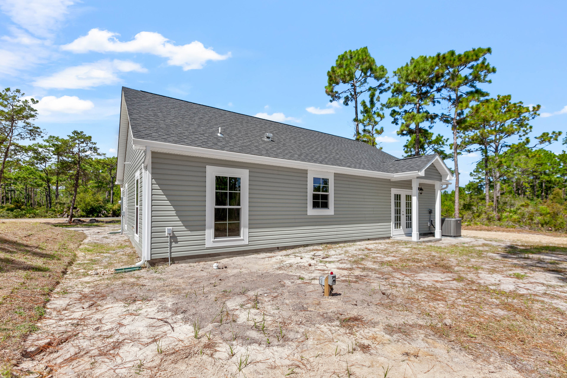 Two-story house with white-framed windows, light-colored siding, and a grassy yard bordered by mature trees; grey utility box sits near the driveway.