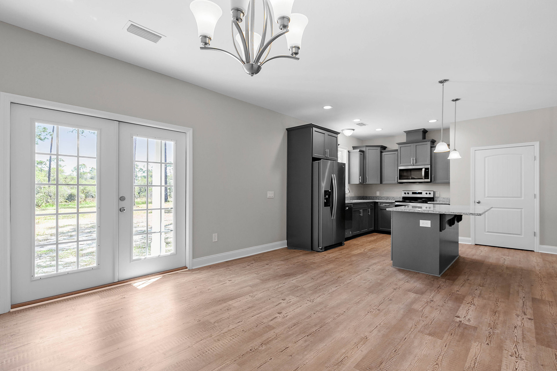 Open kitchen and dining area featuring wood flooring, white cabinetry, stainless steel refrigerator and microwave, glass-paned double doors, and a modern chandelier.