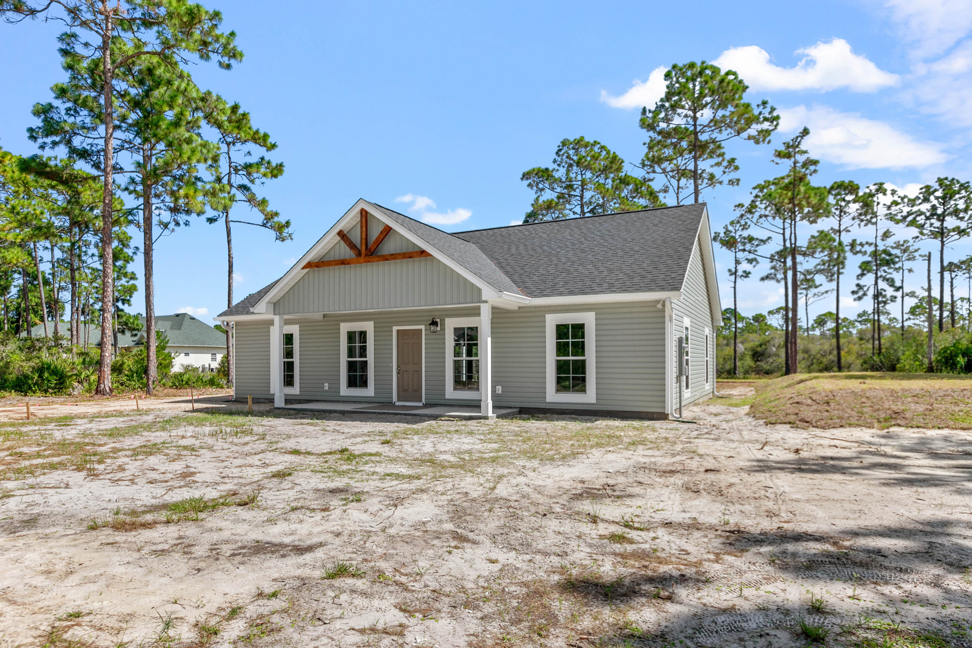Two-story home with gray siding, white trim, covered front porch, large windows, and dirt lot in foreground; mature trees and cloudy sky in background