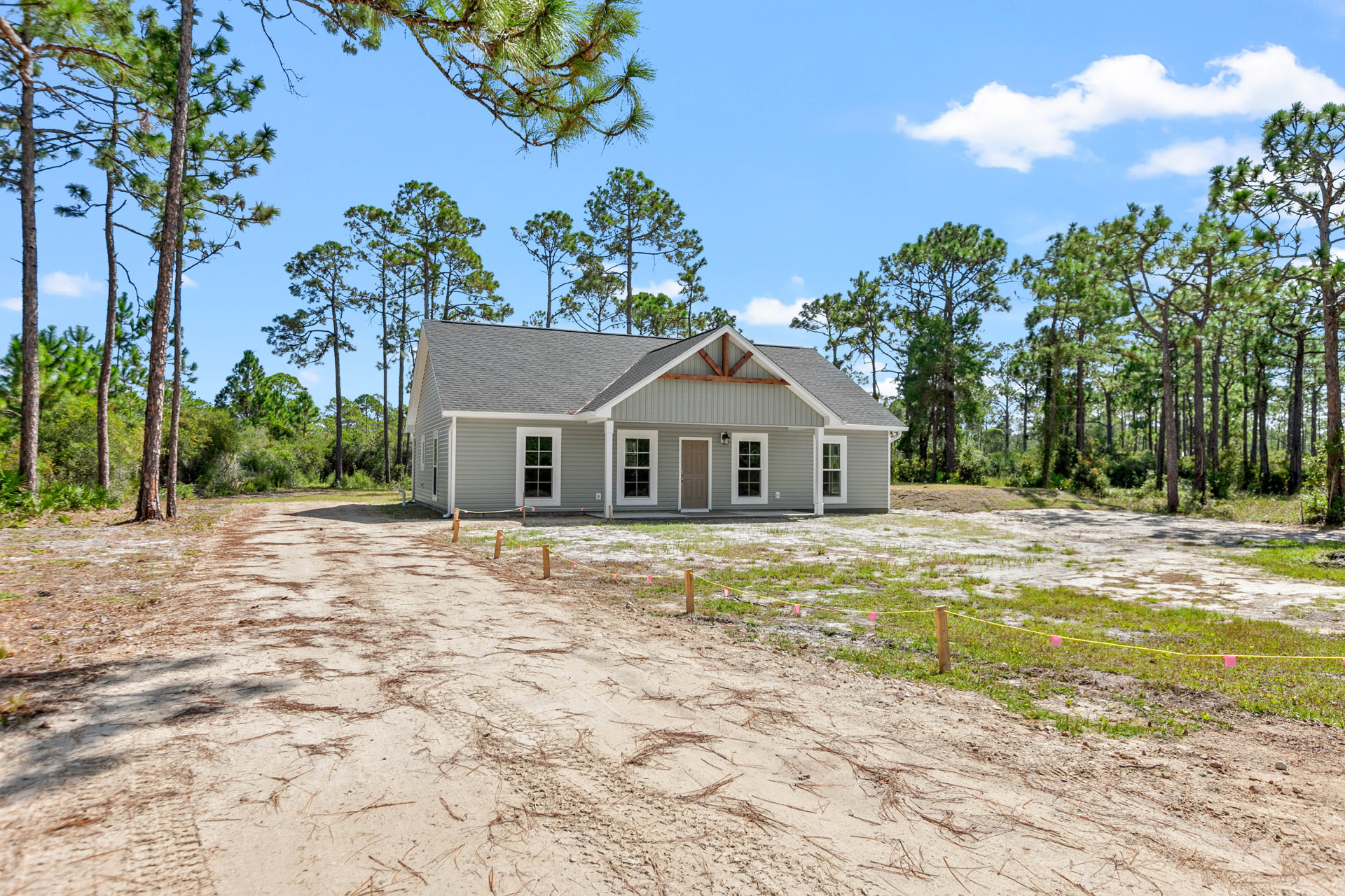Single-story house with light siding, few windows, and gabled roof, set beside a dirt road with tire tracks, grass, and wooden fence; mature trees and cloudy sky in the background