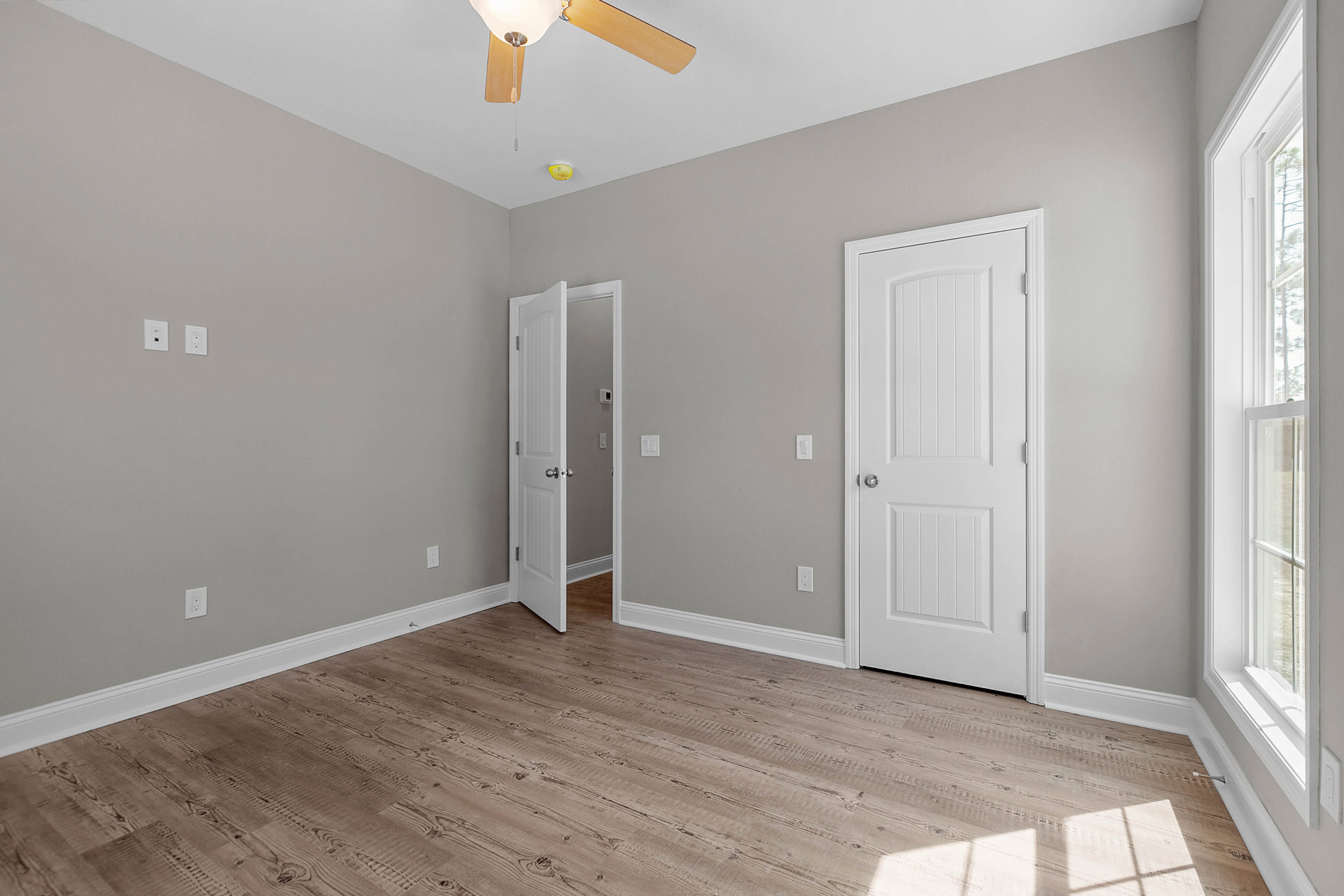 Room with wood laminate flooring, two white doors with silver knobs, ceiling fan, white walls, window with trim, and crown molding.