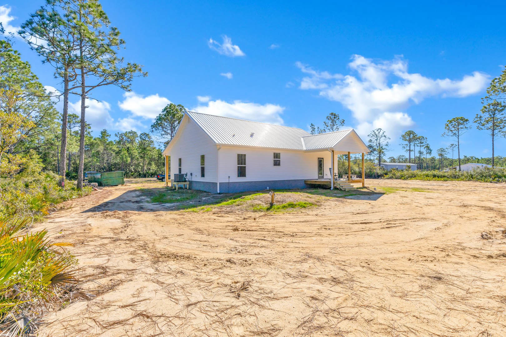 White two-story house with horizontal siding, large windows, and a dirt yard under a clear blue sky, surrounded by tall trees and scattered plants.