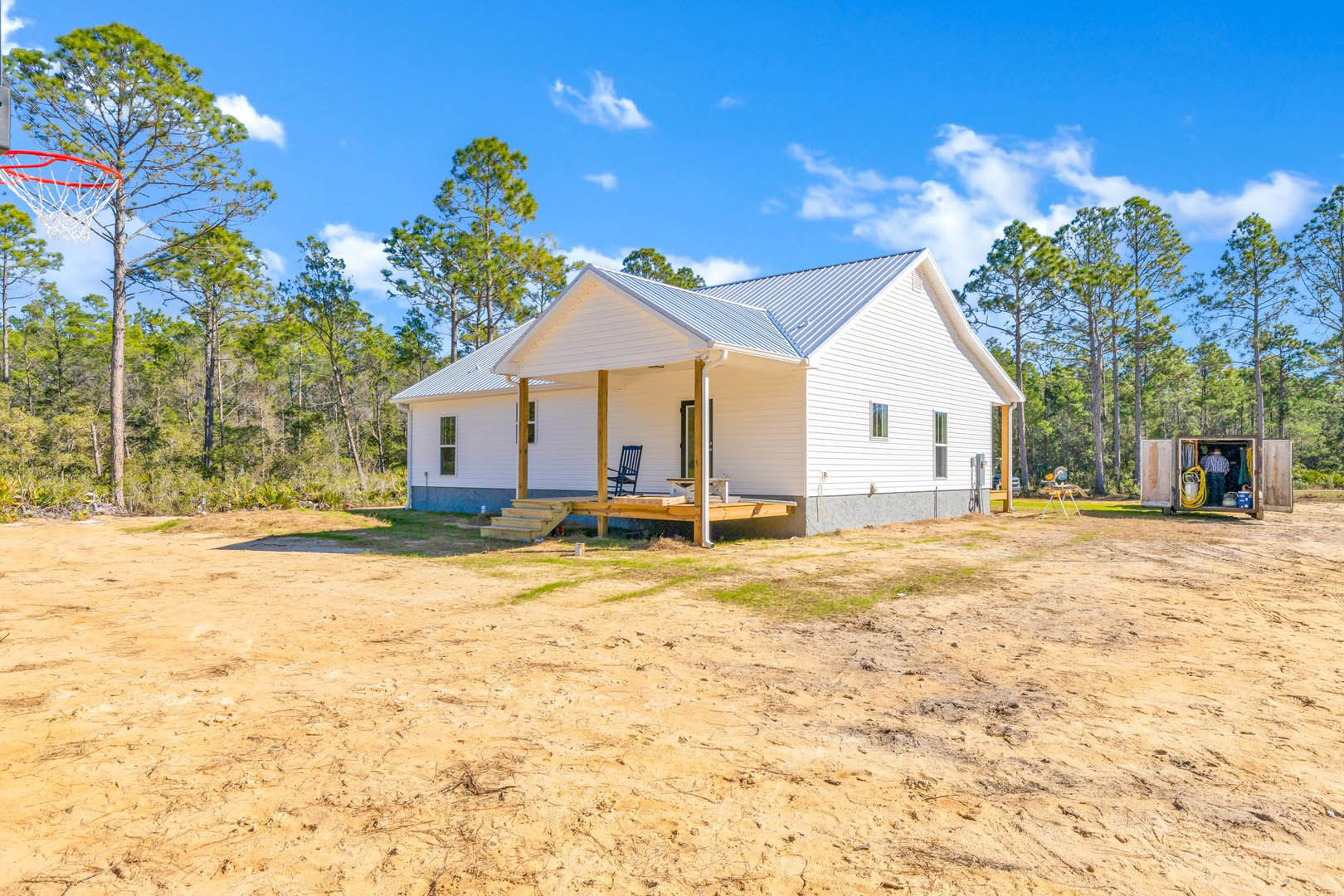 White house with blue roof, front porch, and deck, surrounded by dirt lot and trees in the background