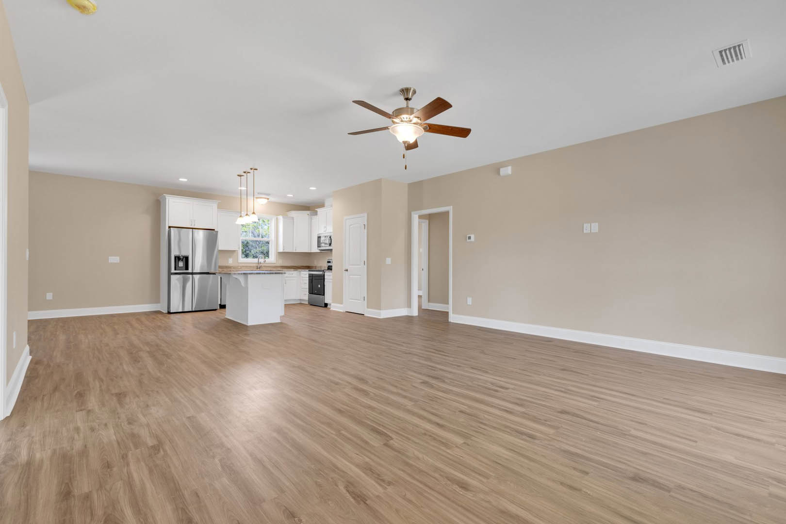 Spacious room featuring hardwood flooring, white walls, ceiling fan with light fixture, white door with silver handle, stainless steel refrigerator, and a person beside a white