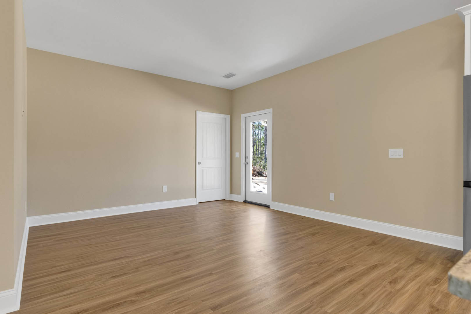 Hardwood floor with white door featuring silver handle, white walls, window showing trees outside, white ceiling with air vent