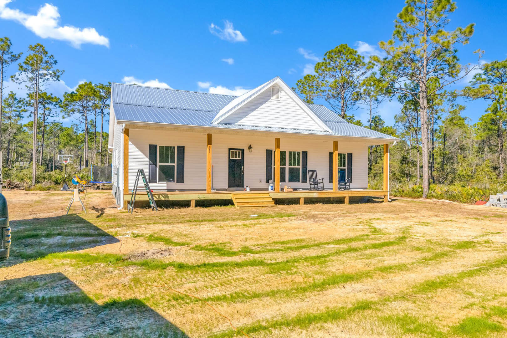 Two-story house with blue roof, white siding, covered porch, grassy yard, wooden fence, mature trees in background, and outdoor chair near entrance