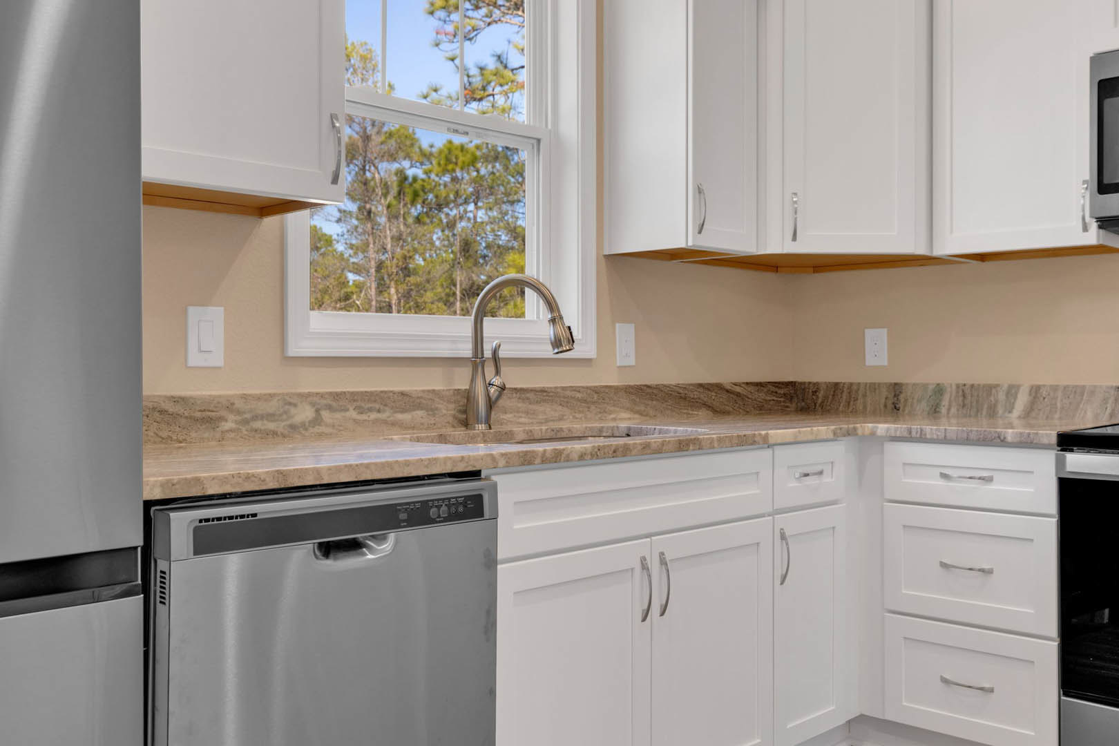 Modern kitchen with white shaker cabinets, stainless steel sink and faucet, built-in dishwasher, light stone countertop, large window overlooking green trees, and sleek white