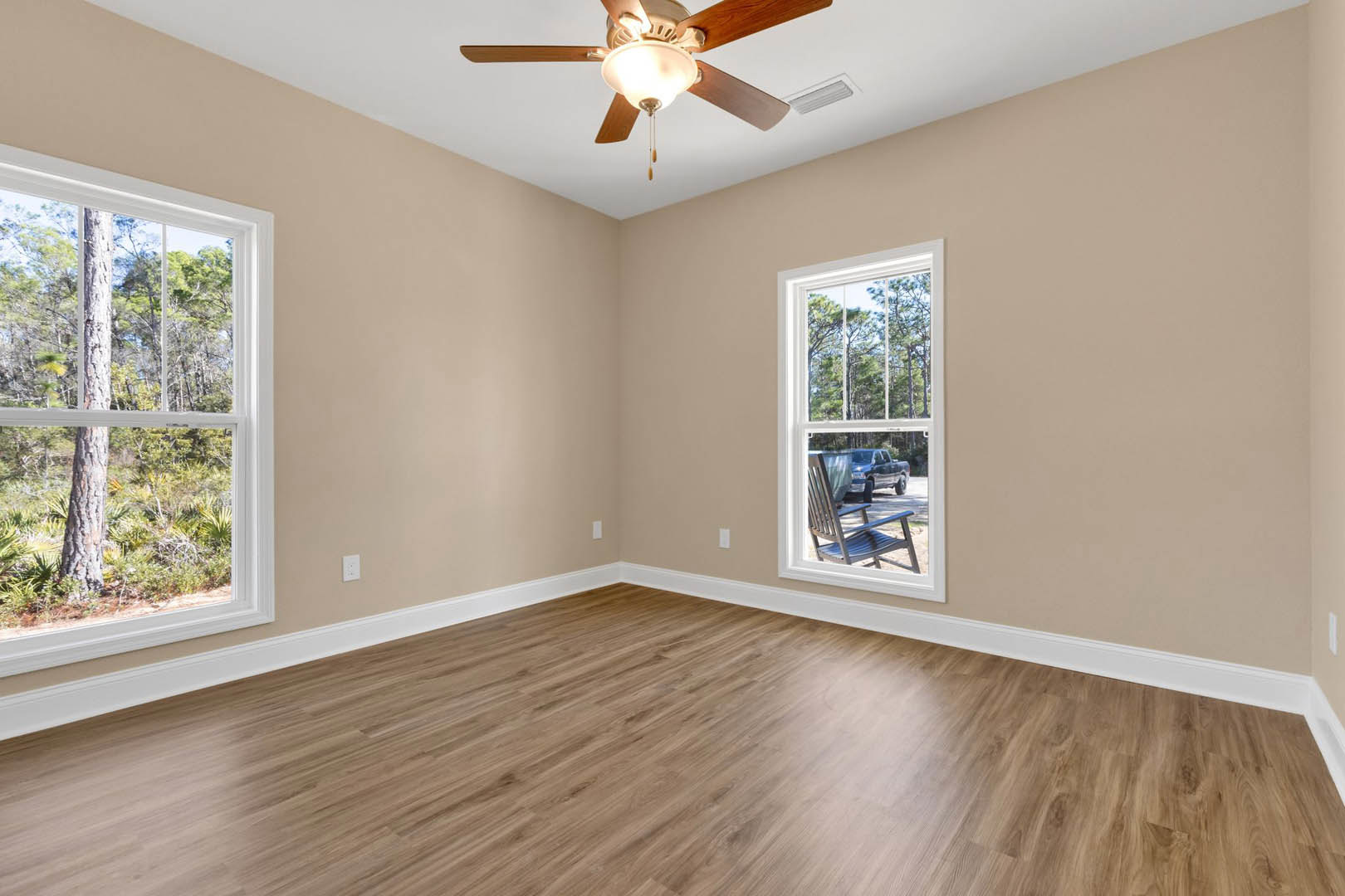 Living room with wood flooring, white trim, ceiling fan with light fixture, large windows showing trees and a parked truck outside