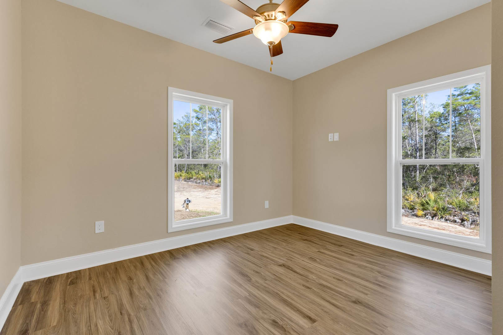 Living room with hardwood flooring, ceiling fan with light fixture, large windows framed by white molding, and outdoor trees visible through glass.