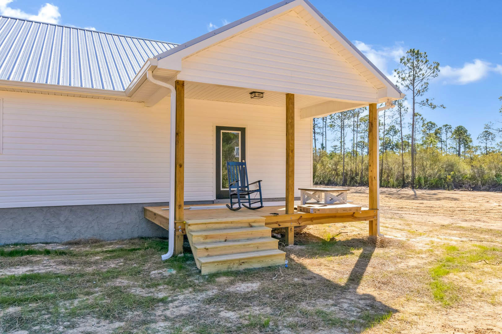 Wooden porch with blue rocking chair beside window, staircase leading to entry, tall leafy tree providing shade, white siding exterior