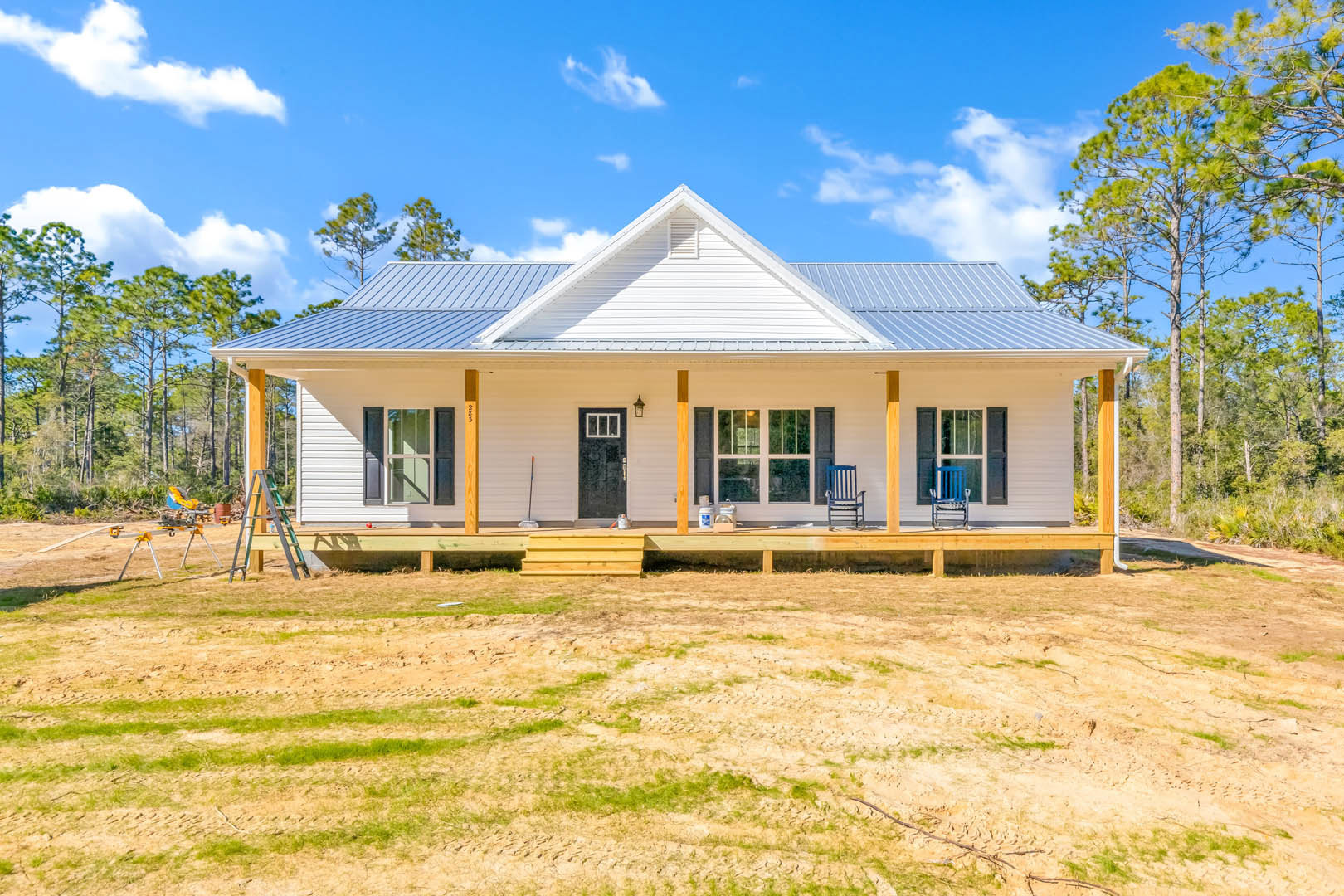 Two-story house with blue metal roof, covered front porch, black door with white trim, blue railing, blue chair, surrounded by dirt field and sparse grass under partly cloudy sky.