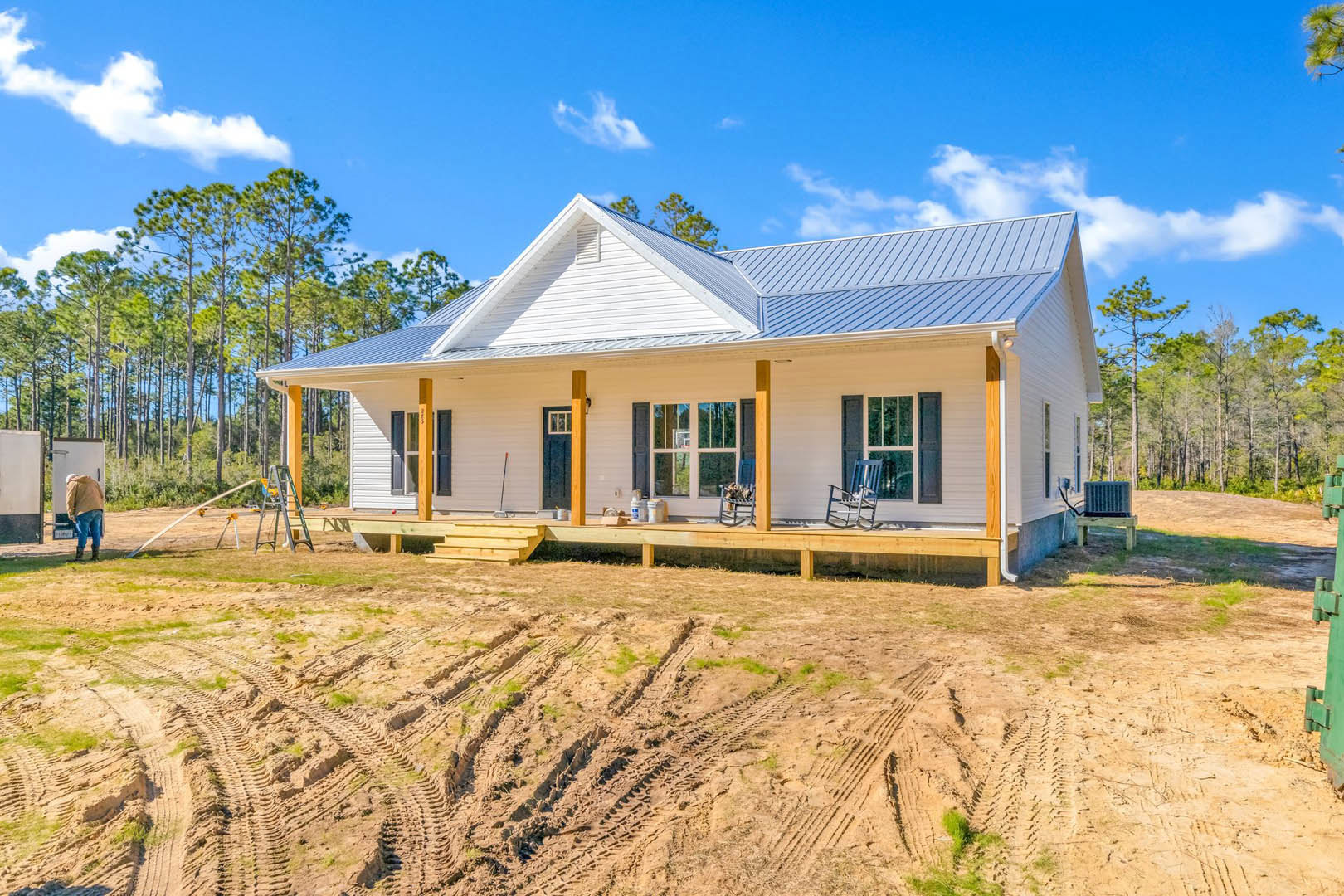 Modern house with blue metal roof, large windows, and dirt yard featuring tire tracks; a person stands near the entrance, blue chair sits on concrete patio, construction equipment