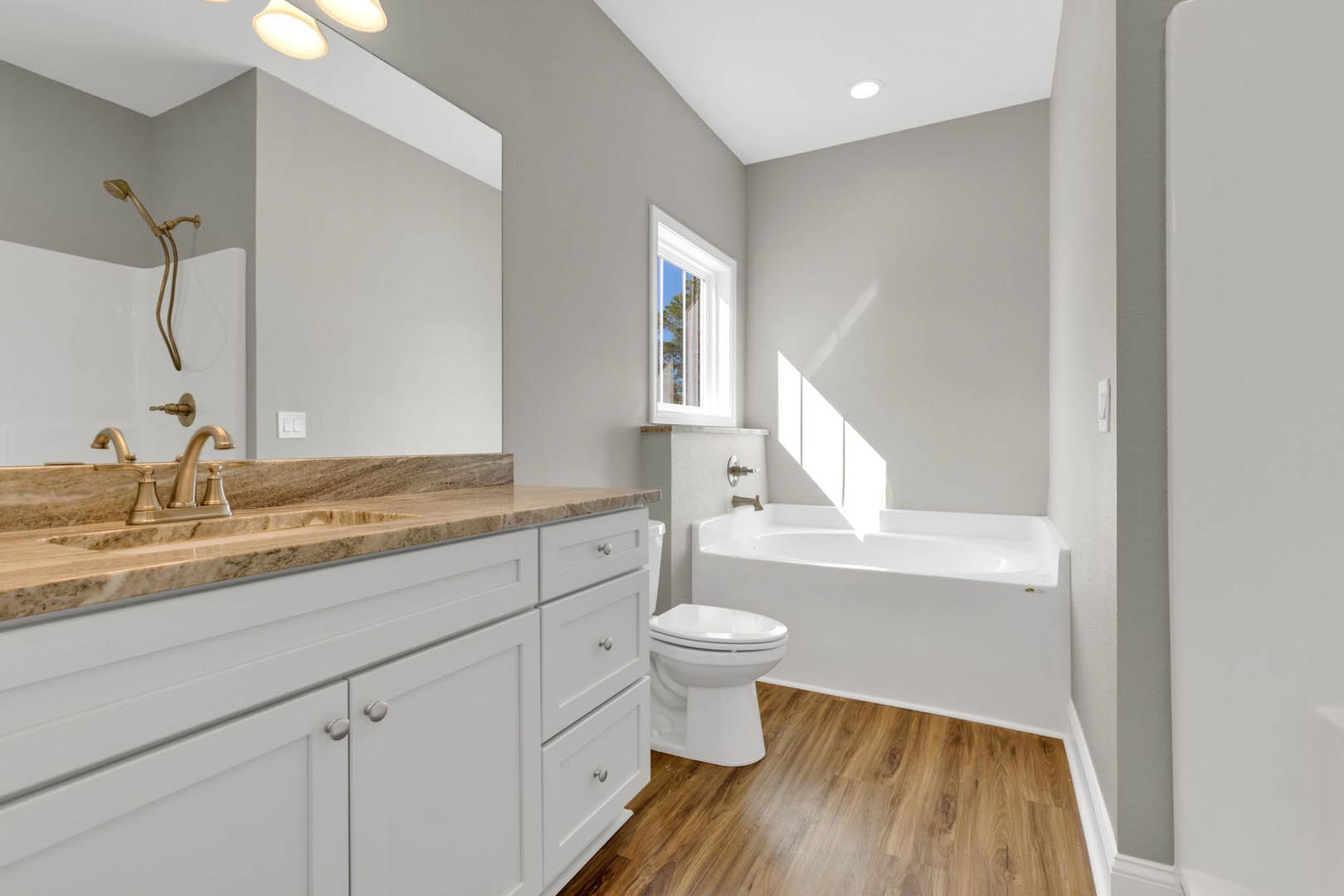 White bathtub and toilet set against light tile walls, chrome shower head with hose, window above tub showing blue sky, white countertop partially visible in foreground.