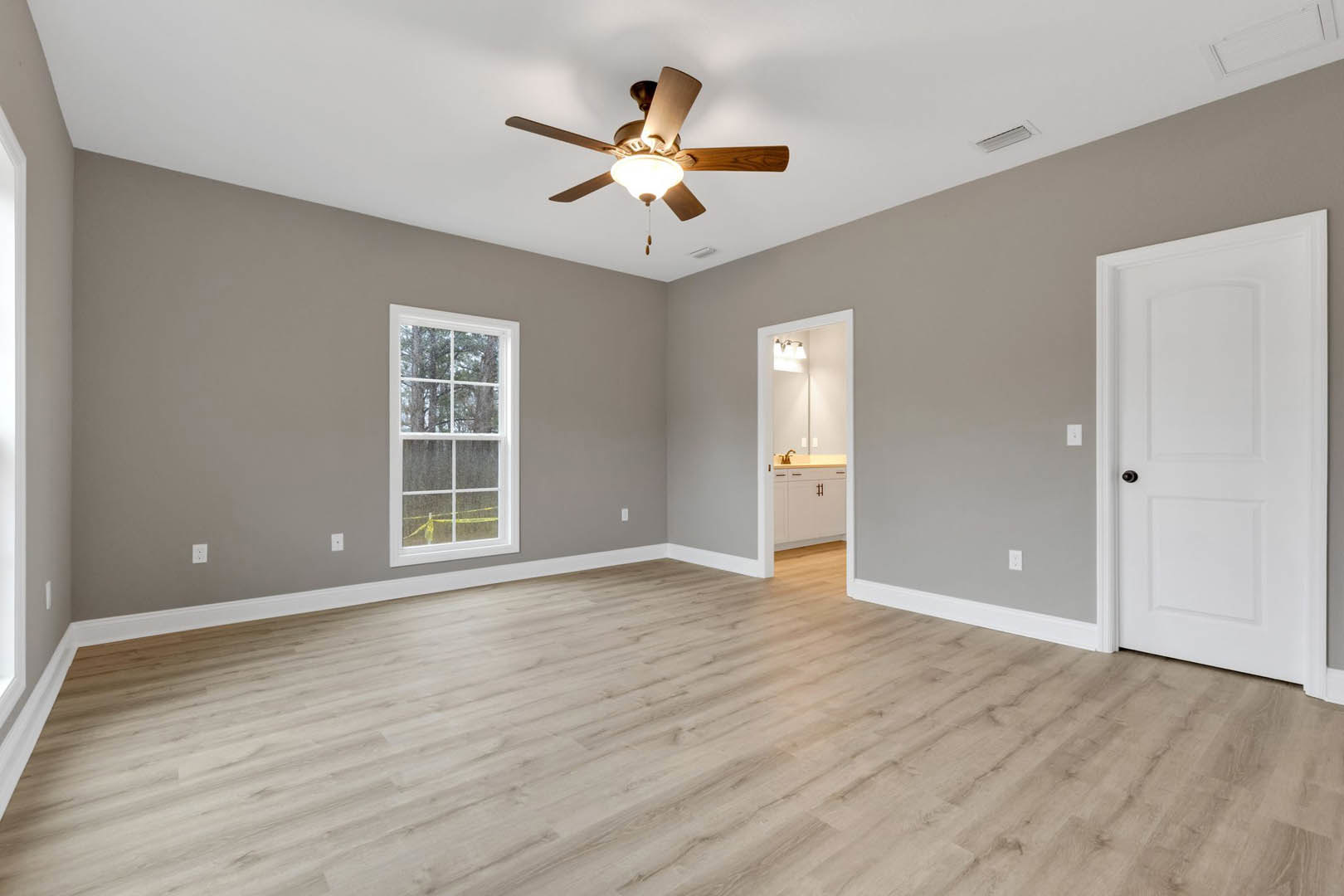 Ceiling fan with light fixture, wood laminate flooring, white walls, window overlooking trees, white door with black handle