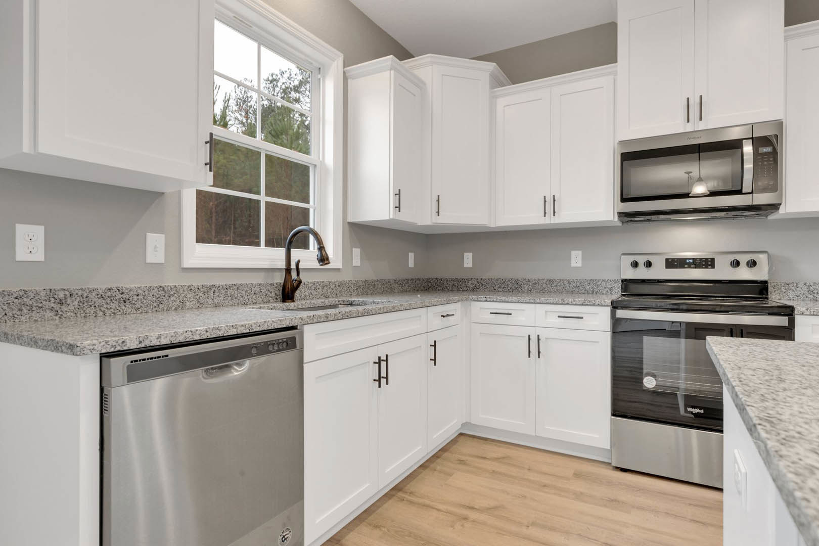 White kitchen cabinets, stainless steel dishwasher beneath quartz countertop, chrome faucet, ceiling light fixture, window overlooking green trees