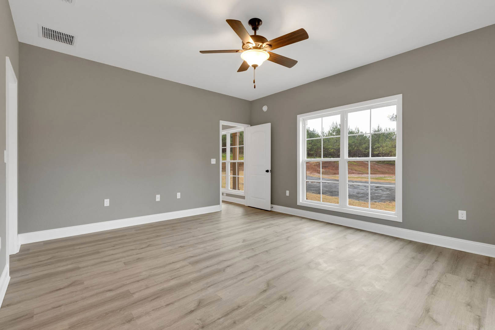 Ceiling fan with light fixture centered above hardwood flooring, large windows framed by white walls, outdoor view of trees, wall vent visible near baseboard.