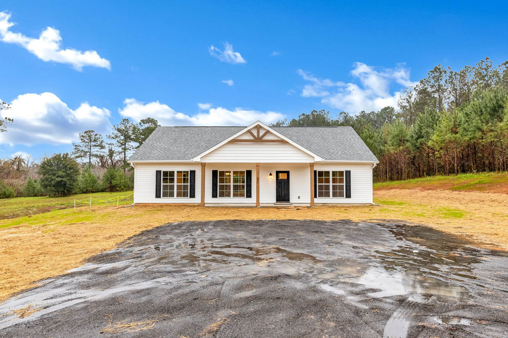 Two-story house with black front door, large windows, gray roof, concrete driveway with puddle, yellow sign near entrance, surrounded by grass and trees under cloudy sky.