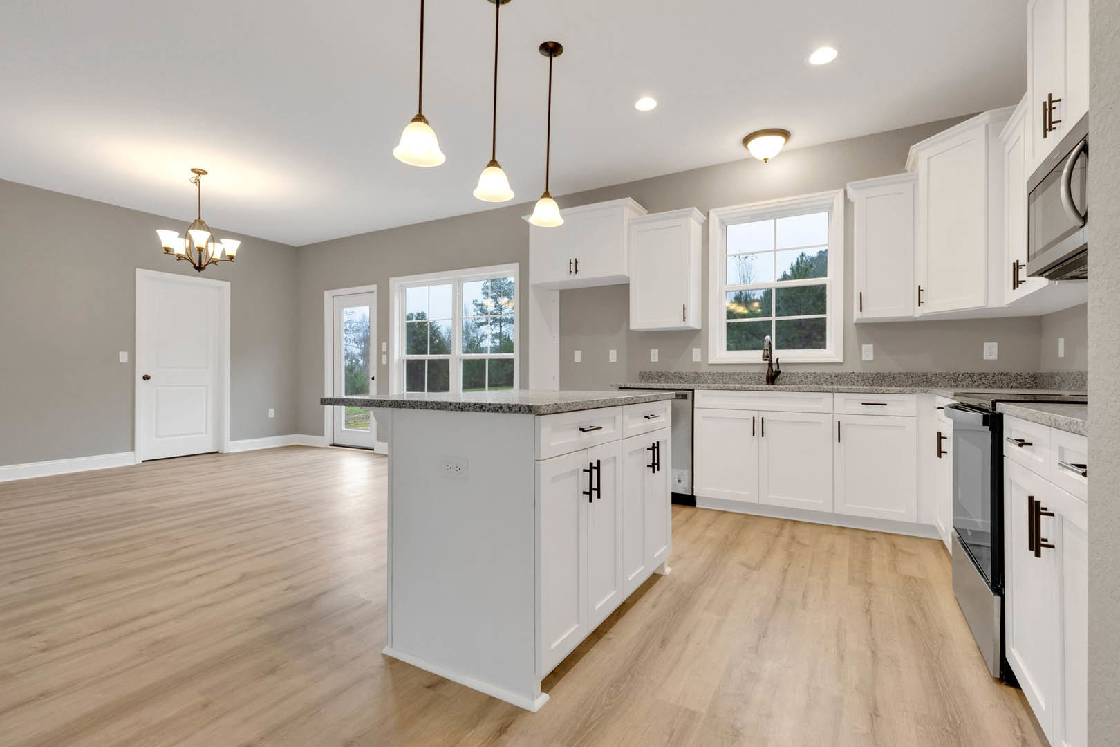 White kitchen cabinets with black handles, wood flooring, ceiling pendant lights, stainless steel appliances, a window overlooking trees, and a white door with a brown handle.