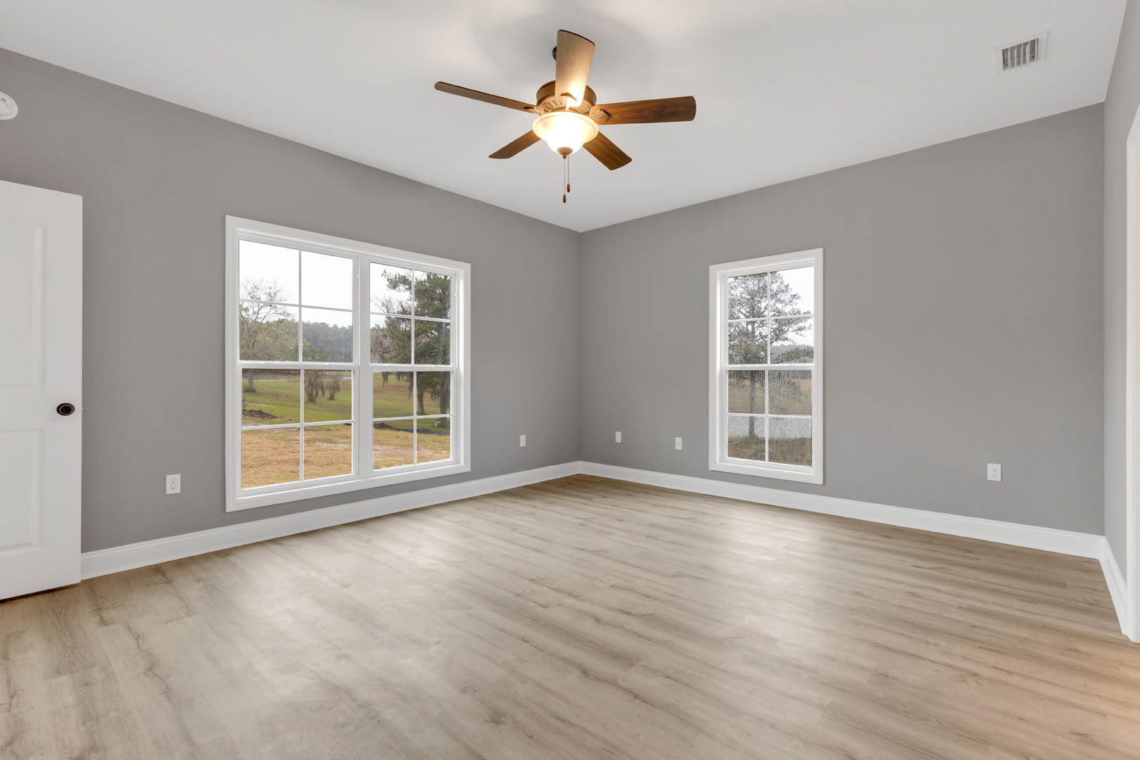 Ceiling fan with light fixture, two large windows showing a rainy field view, wood laminate flooring, white walls, interior door with small hole