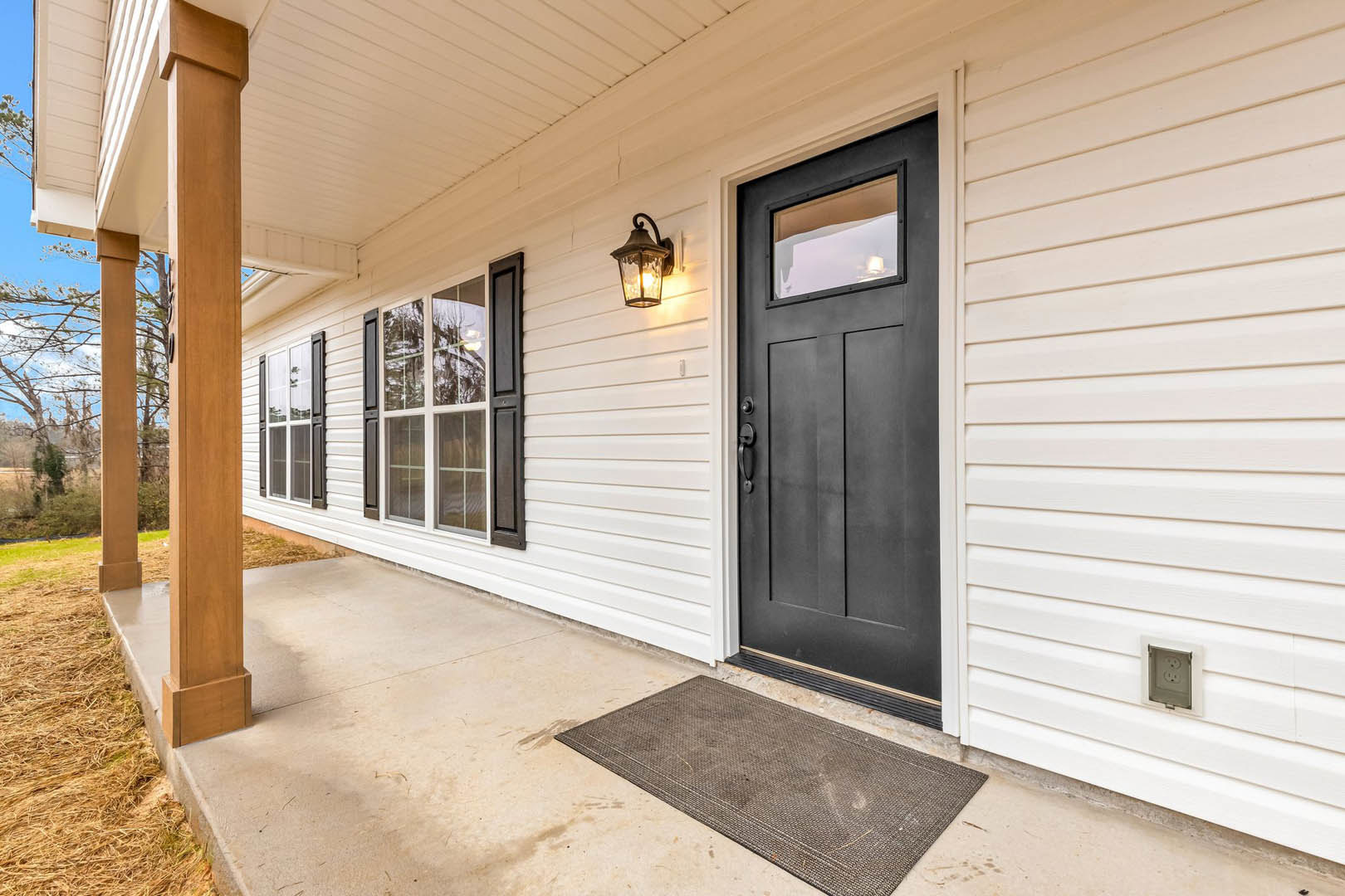 Front porch with wood siding, brown welcome mat, black shuttered window, white door, outdoor light fixture, and electrical outlet