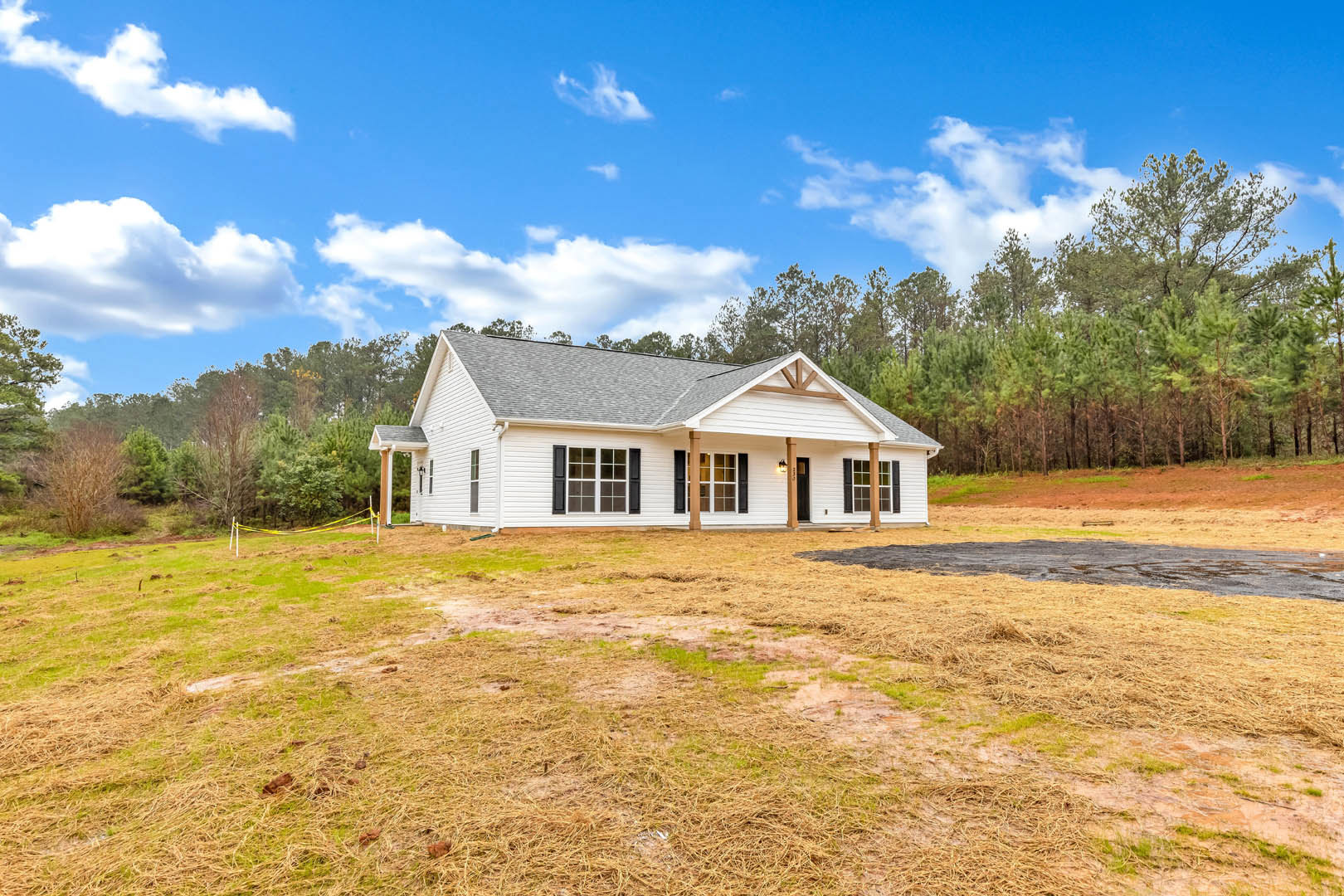 Two-story house with gray siding, covered front porch, large grassy yard, mature trees in background, white-framed windows, gabled roof