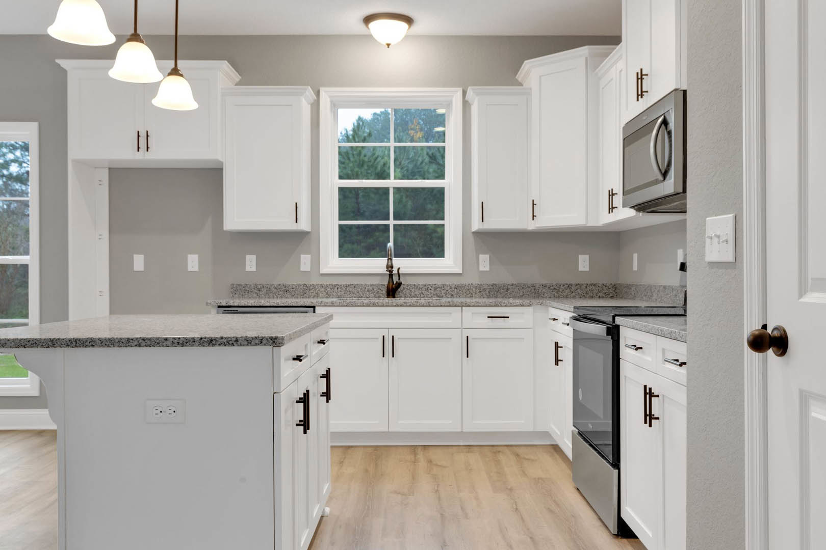 Kitchen with white cabinets, granite countertops, stainless steel microwave, ceiling lights, window overlooking trees, and white wall switch with three knobs