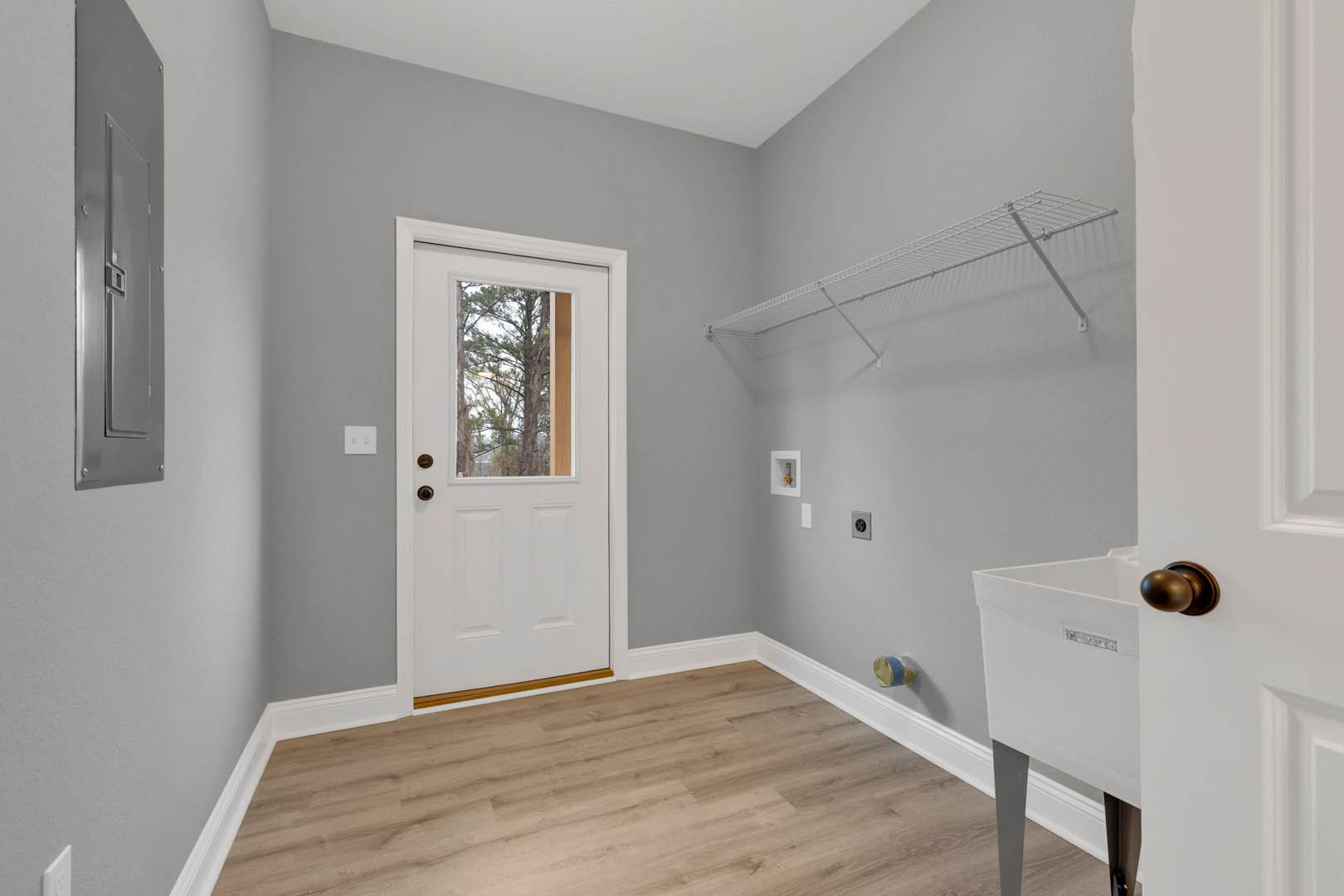 Laundry room with wood flooring, white door featuring a window, white wall-mounted shelf, white utility sink, and window overlooking trees; blue and yellow cylinder placed near the