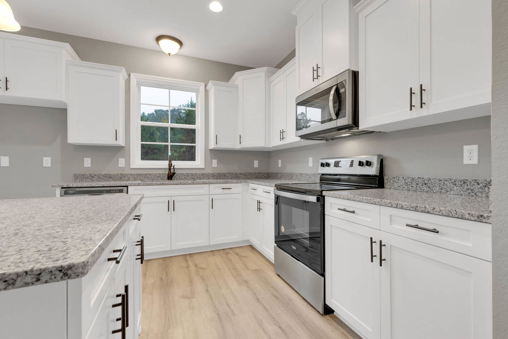 White kitchen cabinets and granite countertops, stainless steel stove and microwave, window overlooking trees, white door with black handle, white electrical outlet with black