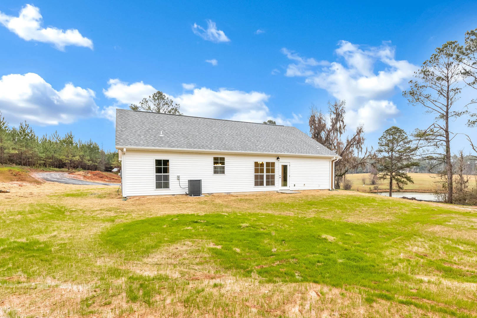 White farmhouse with white-framed windows, air conditioner unit, and pitched roof; green lawn in front, trees and blue sky in background.
