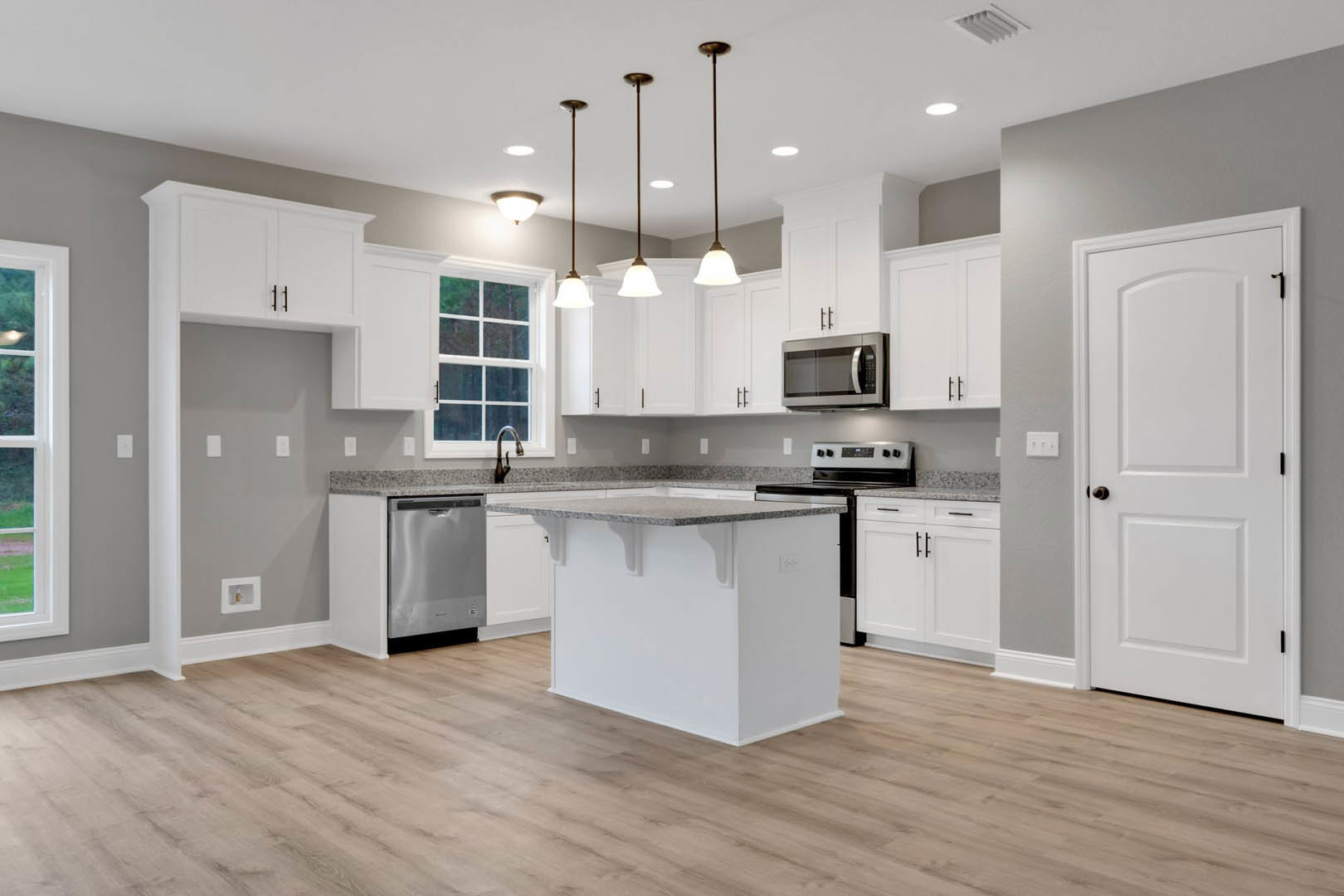 White kitchen with shaker cabinets, granite island countertop, open microwave, black-handled door, close-up of white dishwasher, and decorative bottle in square wall niche.