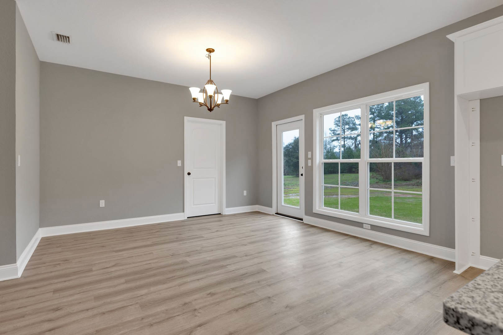 Hardwood floor room with white walls, black door knob on white door, large windows overlooking grassy field, modern chandelier, marble countertop