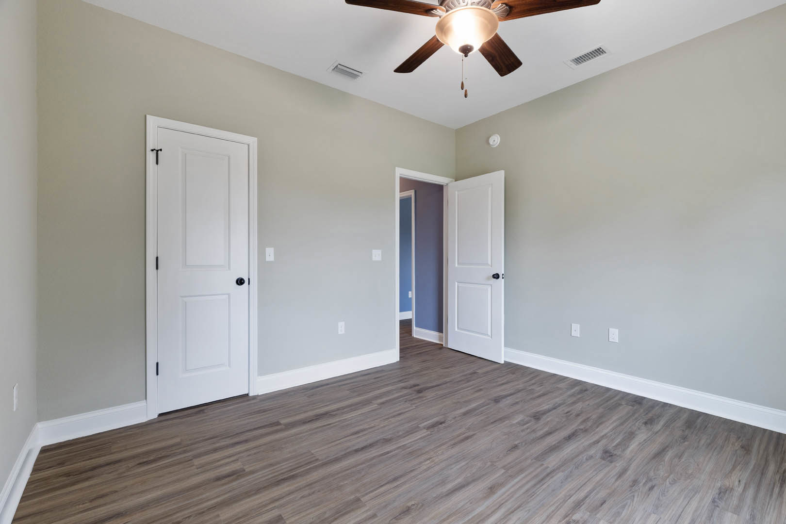 Wood flooring with white baseboards, white doors featuring black knobs, ceiling fan with light fixture, and ceiling vent in a neutral-toned room.