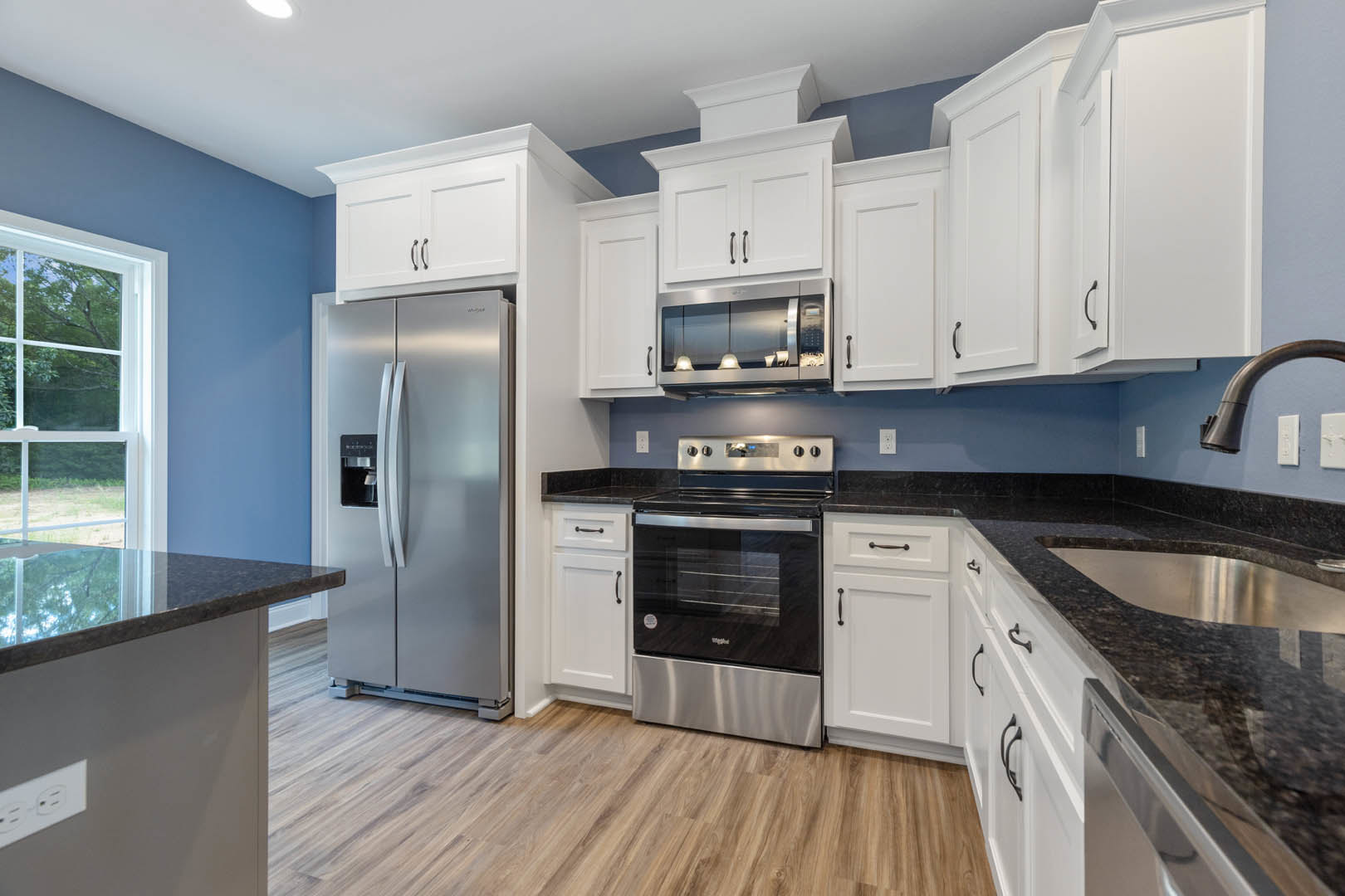 White kitchen cabinets with black countertops, stainless steel stove and refrigerator, undermount sink beneath a window, modern appliances and sleek cabinetry