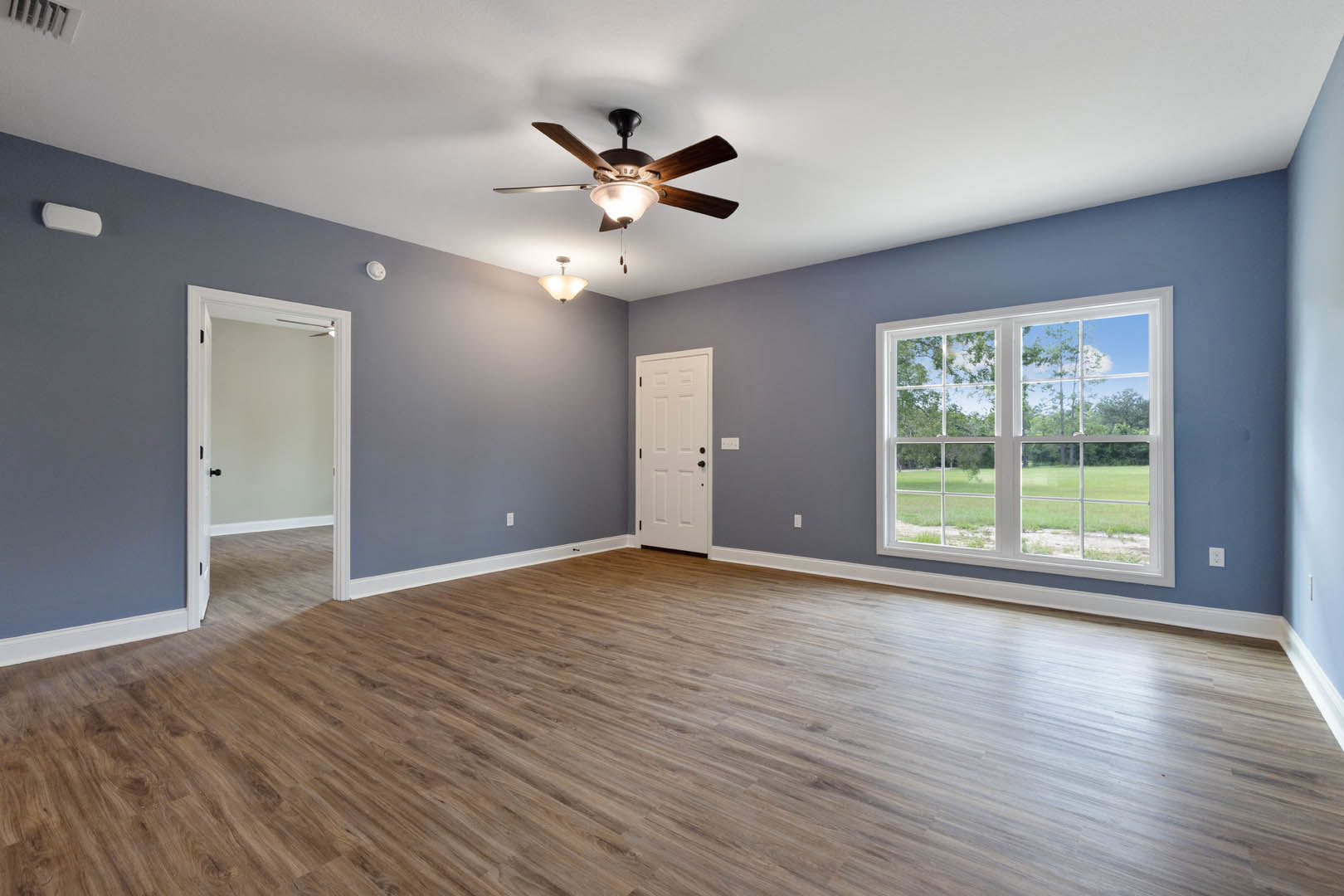 Ceiling fan with light fixture, hardwood floor, white door with black handle, window overlooking grassy field, neutral walls and trim