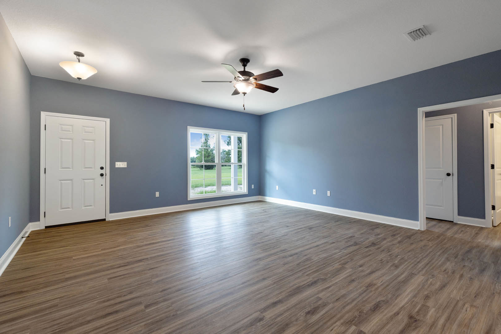 Hardwood floor room with white door featuring black handle, ceiling fan with light, and window overlooking green lawn
