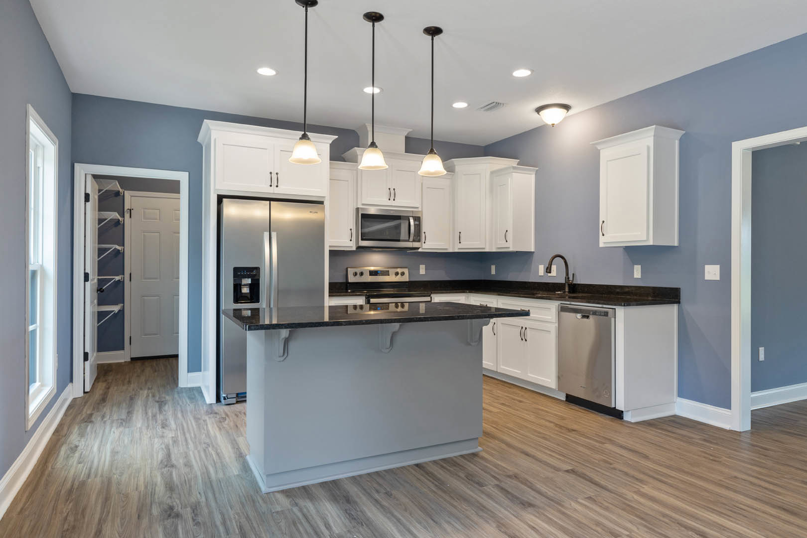 White kitchen cabinets with black countertops, stainless steel microwave, white door with built-in shelves, recessed ceiling lighting, and light-colored flooring.