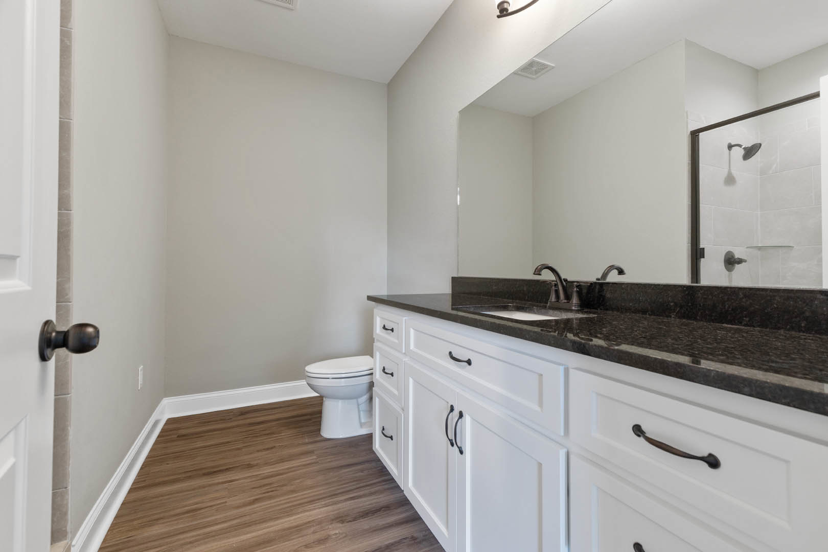 Bathroom with black countertop, large wall mirror, white toilet, wood flooring, tiled shower with chrome showerhead, white cabinetry, and modern faucet.