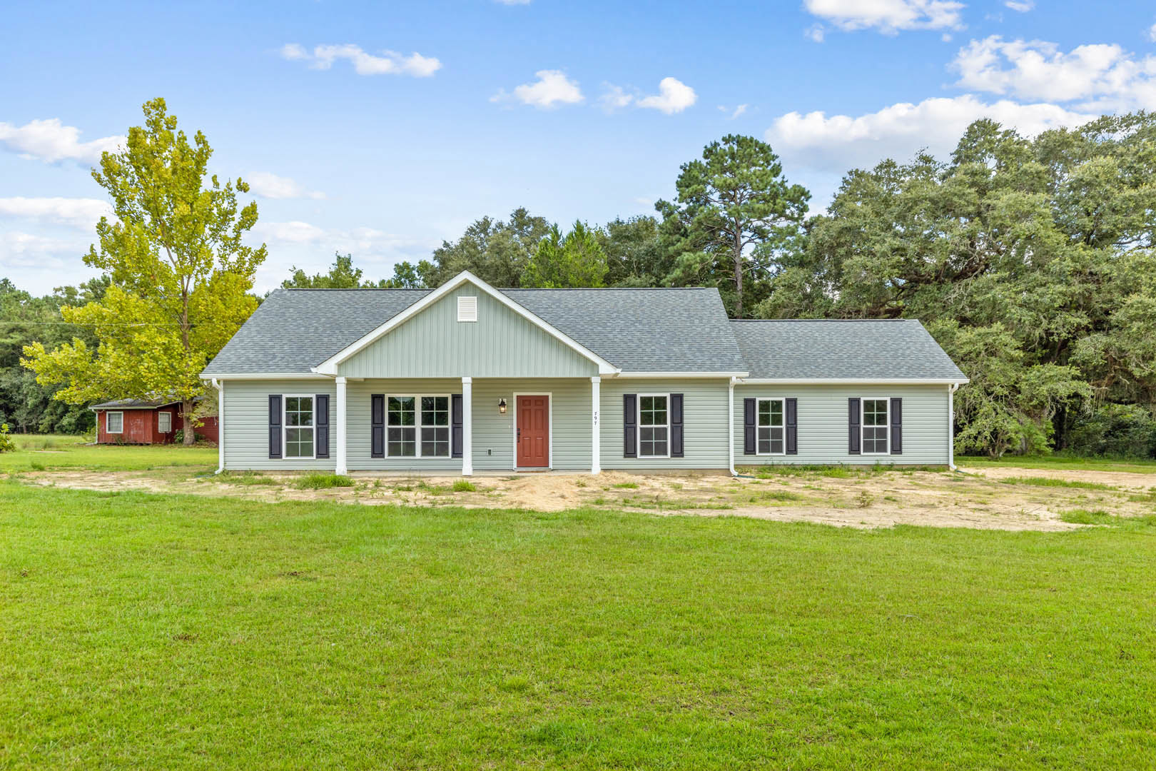 Two-story farmhouse with white siding, black shuttered windows, red front door, manicured lawn, mature trees, and cloudy sky in background