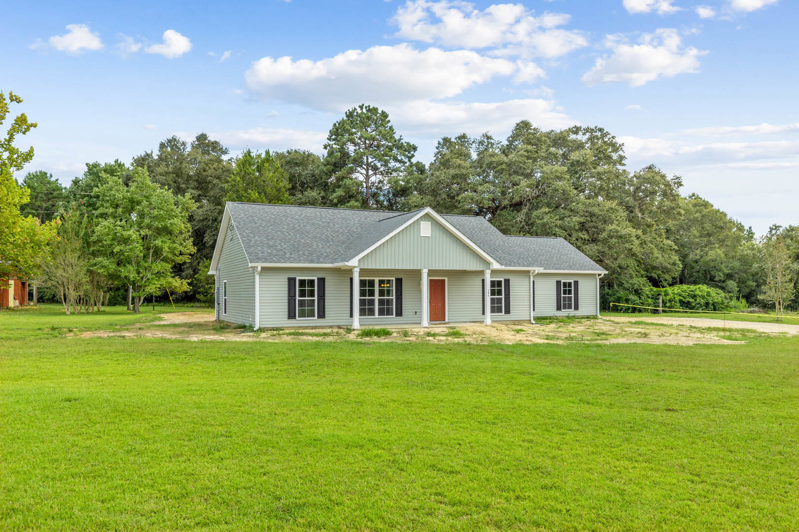 Two-story farmhouse with red front door, multi-pane windows, surrounded by green lawn, mature trees, and blue sky with scattered clouds