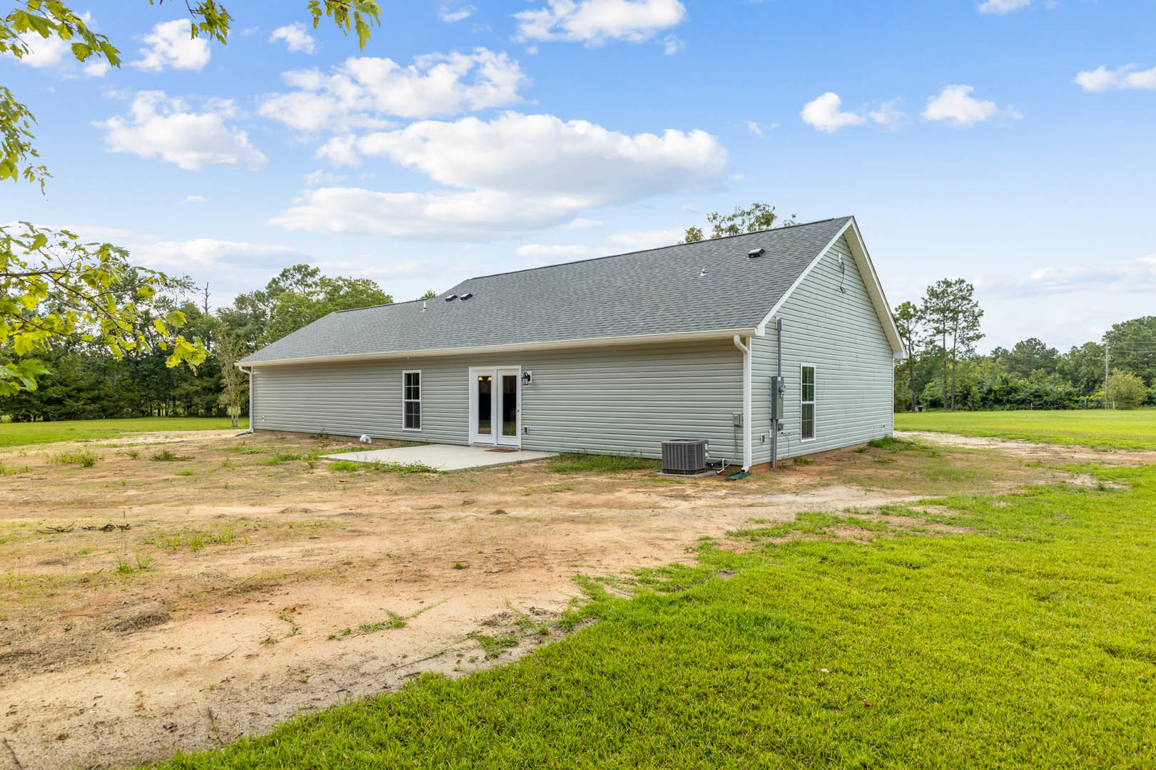 Modern cottage-style home with grey siding, covered porch, white trim, and large windows, set on a dirt lot with scattered trees and blue sky in the background