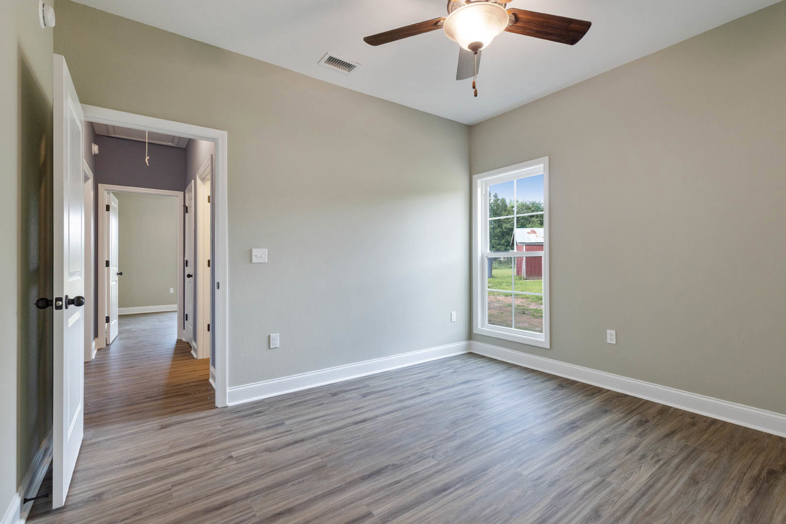 Wood flooring in a spacious room with a ceiling fan and light fixture, large window framing a view of a red barn, neutral walls, and white trim.